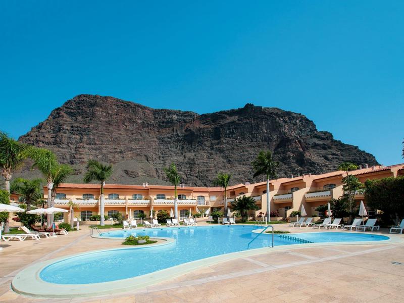 Hôtel avec piscine extérieure et chaises longues devant une montagne rocheuse sous un ciel bleu clair.