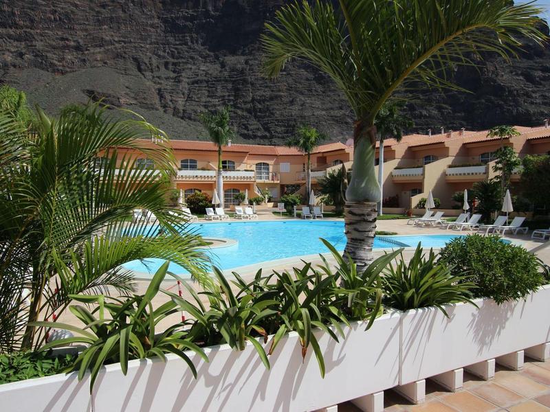 Zone de piscine avec chaises longues et plantes tropicales devant des bâtiments sous des falaises.