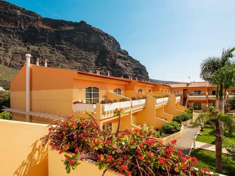 Bâtiments d'hôtel orange devant une falaise rocheuse avec végétation luxuriante et ciel bleu.