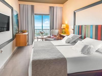 Modernes Hotelzimmer mit Doppelbett, Fernseher, Sessel und Balkon mit Meerblick.