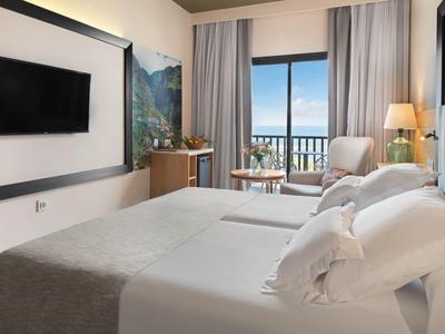 Modern hotel room with two beds, TV, and balcony with sea view.