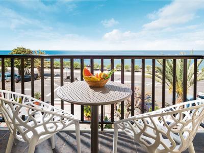 Balcony with two white chairs and a small round table with fruit bowl, view of street and beach.
