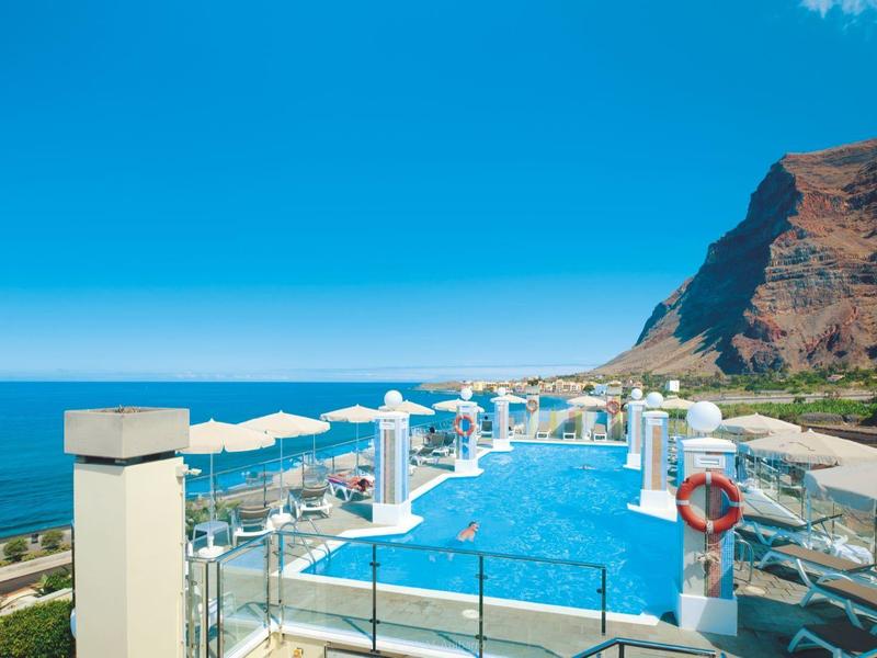Infinity pool with white loungers and umbrellas by cliffs and sea under a blue sky.