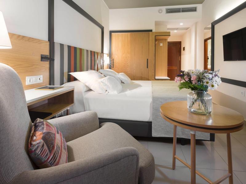 Modern hotel room with comfortable bed, armchair, table, and wall-mounted TV.