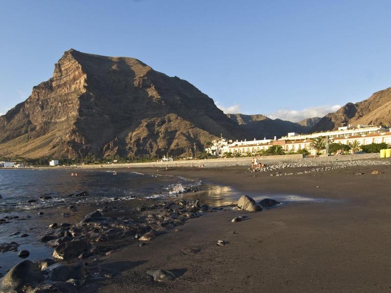 Black sand beach with rocky mountain and some buildings by the sea under clear sky.