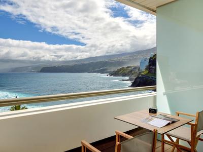 Balkon mit Tisch und zwei Stühlen, Blick auf Meer, Felsen und bewölkten Himmel am Tag.
