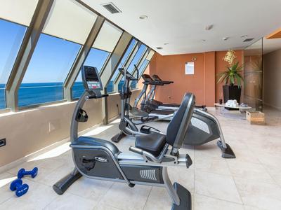 Fitnessraum mit großen Fenstern, Meerblick, Cardiogeräten und blauem Kurzhantel-Set auf hellem Boden.