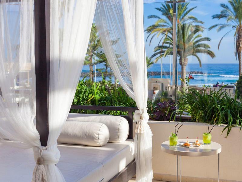 Buiten zitgedeelte met witte gordijnen, kussens en een tafel met drankjes met uitzicht op palmbomen en zee.
