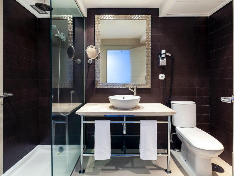 Moderne badkamer met glazen douche, witte wastafel, grote spiegel en toilet op donkere betegelde muren.