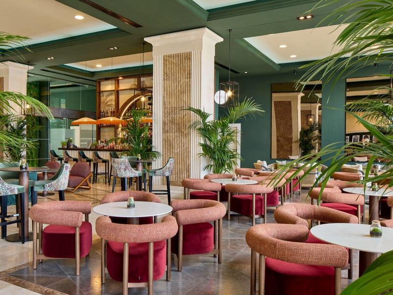 Luxe hotel lounge met roze fluwelen stoelen, ronde tafels en elegante groene planten.