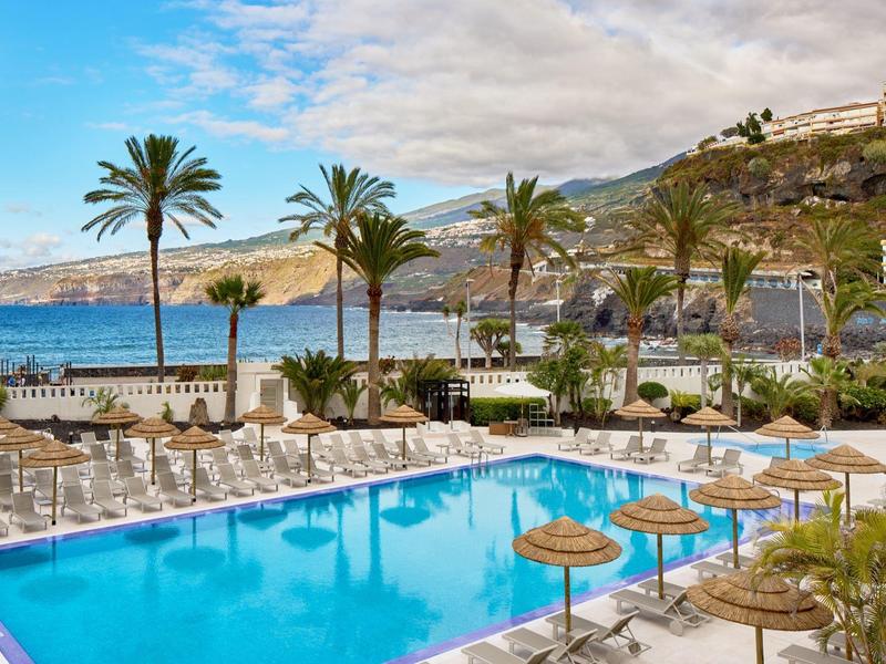 Buitenzwembad met palmbomen, ligstoelen, stroparasols en uitzicht op de oceaan.