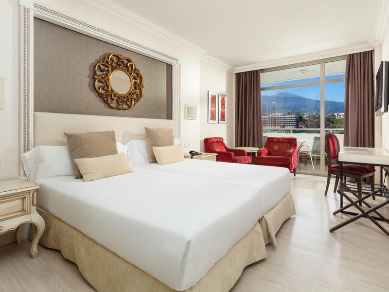 Lichte hotelkamer met tweepersoonsbed, comfortabele stoelen en balkon met uitzicht op bergen.