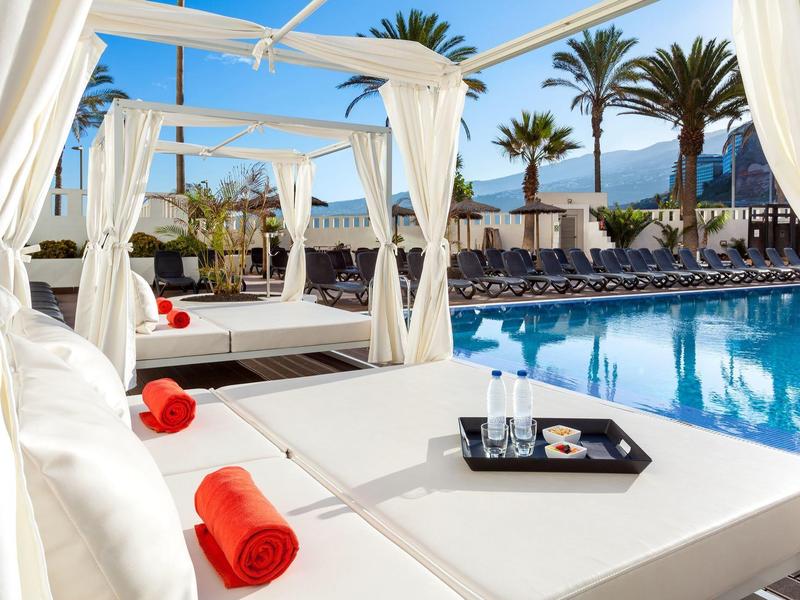 Luxueuze loungestoelen met rode handdoeken naast een zwembad met palmbomen en uitzicht op de bergen.