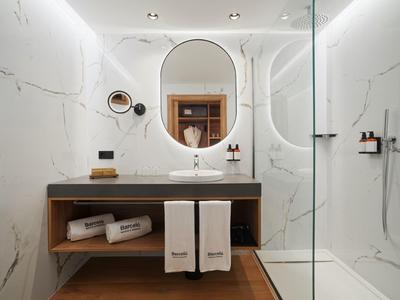 Modernes Badezimmer mit Holzwaschtisch, ovalem Spiegel und begehbarer Glasdusche mit weißen Marmorfliesen.