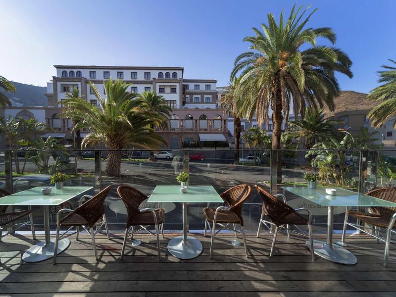 Terraza vacía con mesas y sillas junto a palmeras y un gran edificio al fondo.