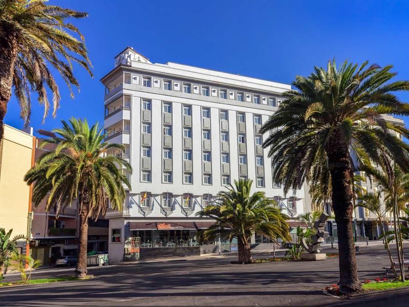 Un edificio blanco de varios pisos con palmeras bajo un cielo azul y una calle ancha.