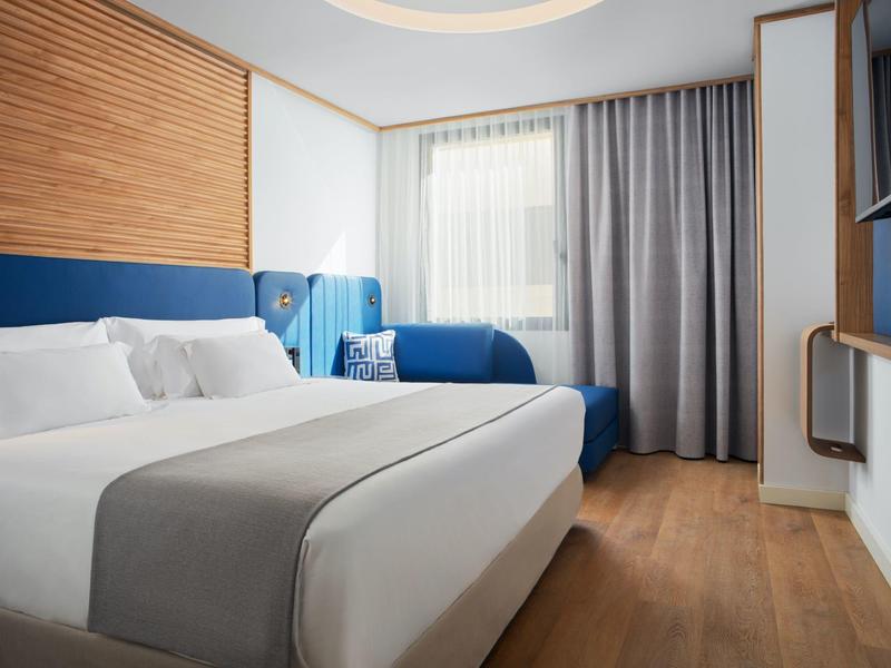 Helles Hotelzimmer mit großem Bett, blauem Sofa und Vorhängen am Fenster