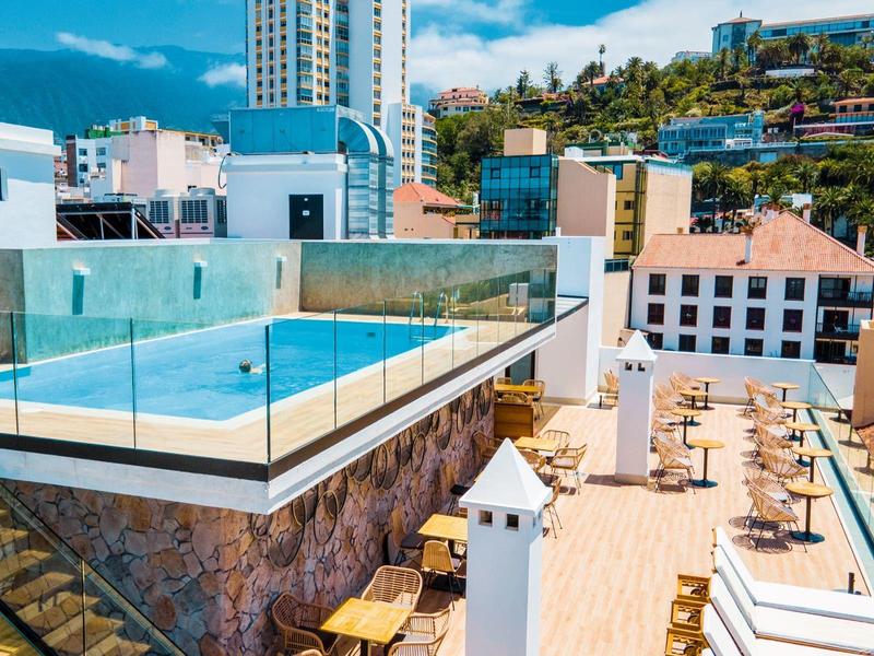 Une piscine d'hôtel moderne sur une terrasse avec une balustrade en verre offrant une vue sur les bâtiments environnants.