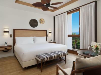 Camera d'albergo luminosa con letto grande, finestra, ventilatore a soffitto e area salotto.