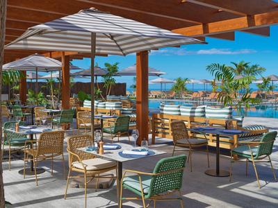 Buitenrestaurant met tafels, stoelen en parasols naast een zwembad onder een blauwe lucht.
