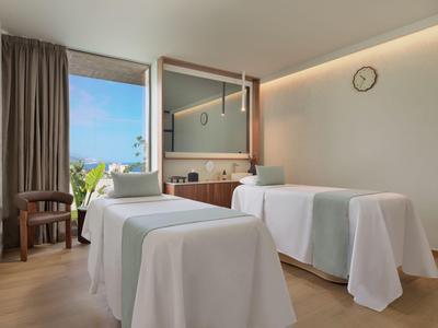 Moderne hotel spa kamer met twee witte massagetafels, uitzicht op terras en planten.