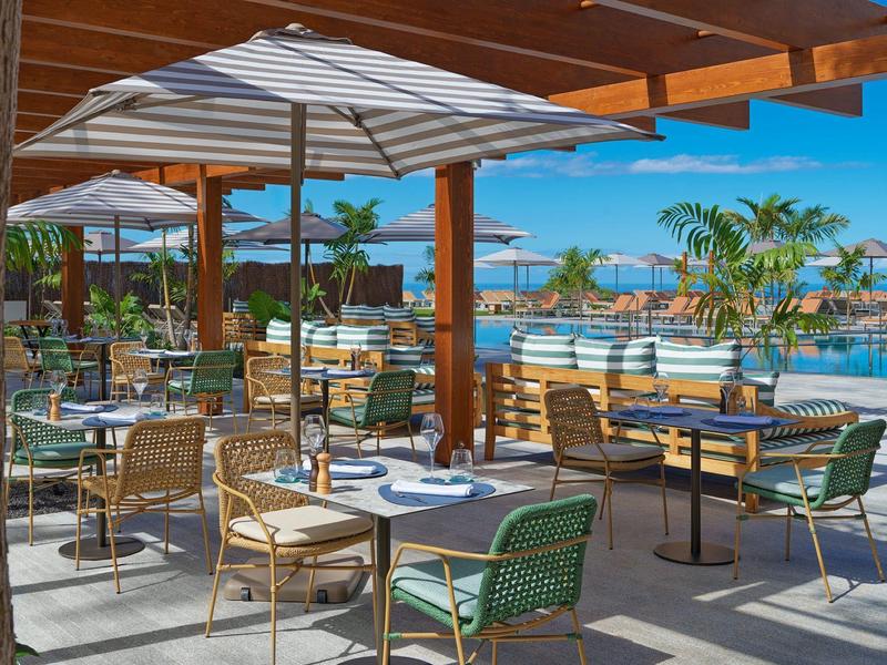 Ristorante all'aperto con ombrelloni e vista sulla piscina e sul mare sotto un cielo limpido.