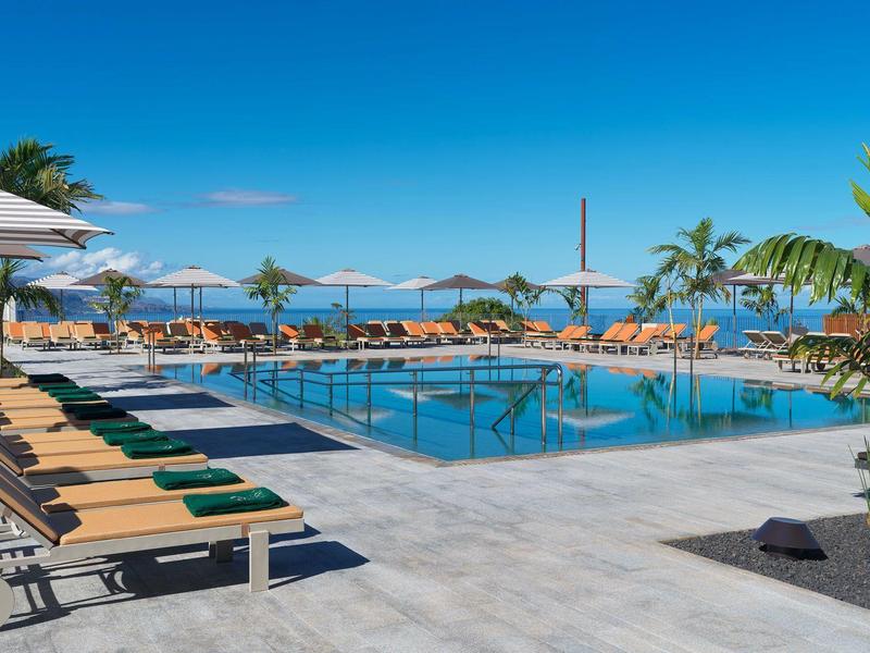 Area piscina di un hotel moderno con lettini, palme e vista sul mare sotto un cielo blu.