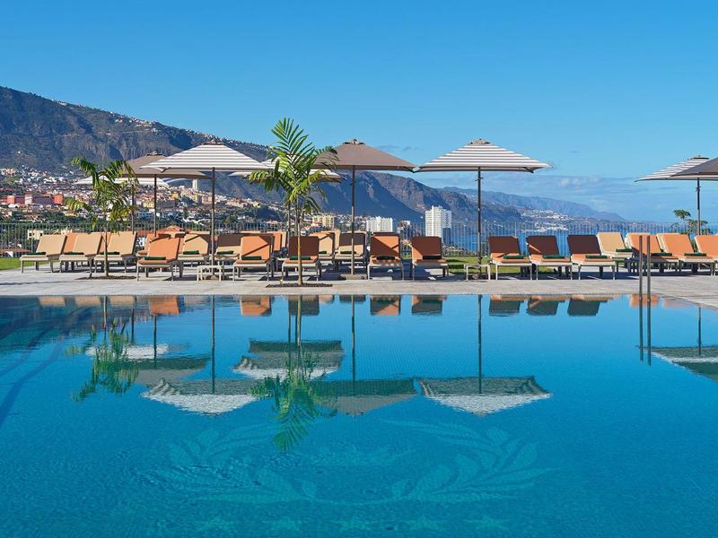 Piscina a sfioro con lettini e ombrelloni davanti a montagna e cielo sereno