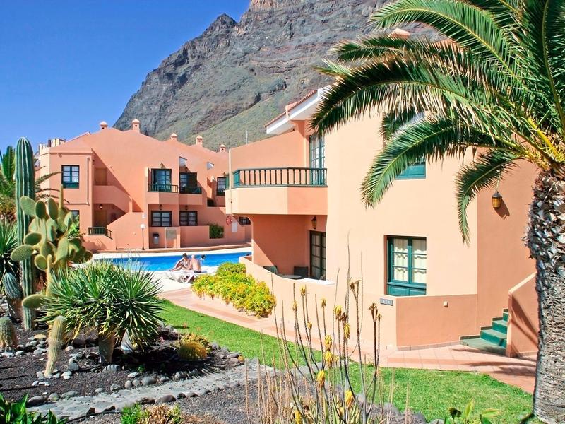 Hotelové budovy s bazénem a palmami před skalní stěnou za jasné oblohy.