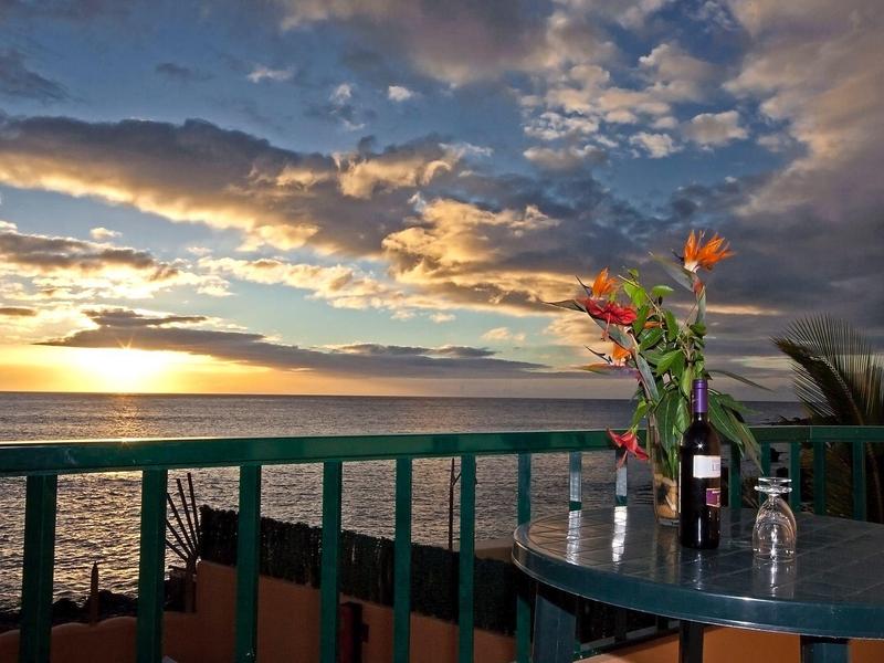 Výhled na moře při západu slunce se stolem, květinami a sklenicí vína na balkóně.