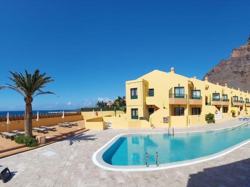 Žlutý hotel s bazénem, výhledem na moře a palmami před skalnatou horou pod modrou oblohou.