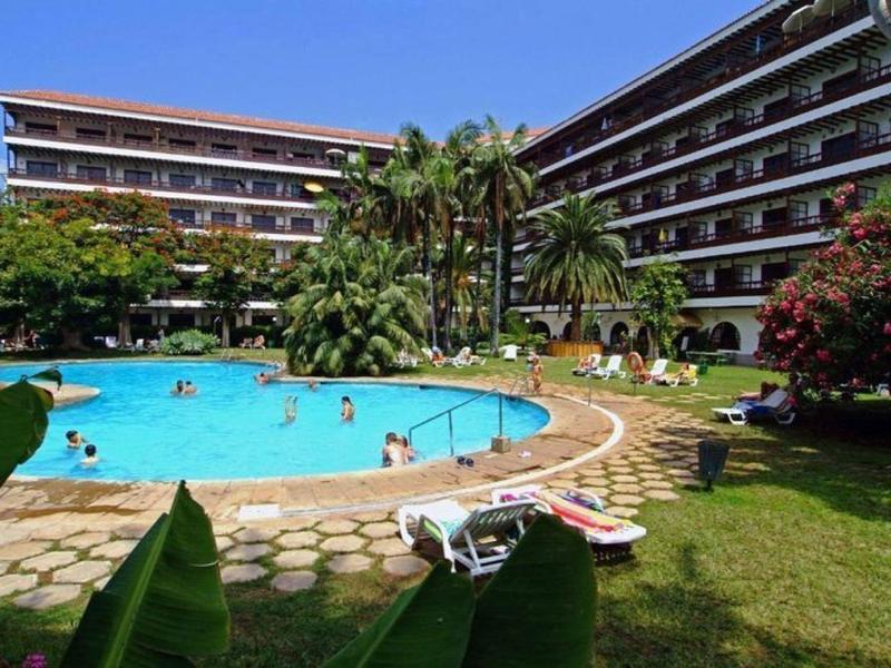 Grande hotel con piscina curva, palme e bagnanti su prato soleggiato.