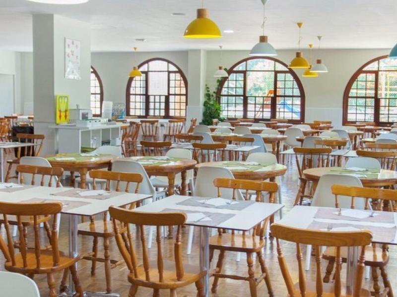 Ampia sala da pranzo luminosa con sedie in legno e tavoli bianchi sotto lampade sospese.