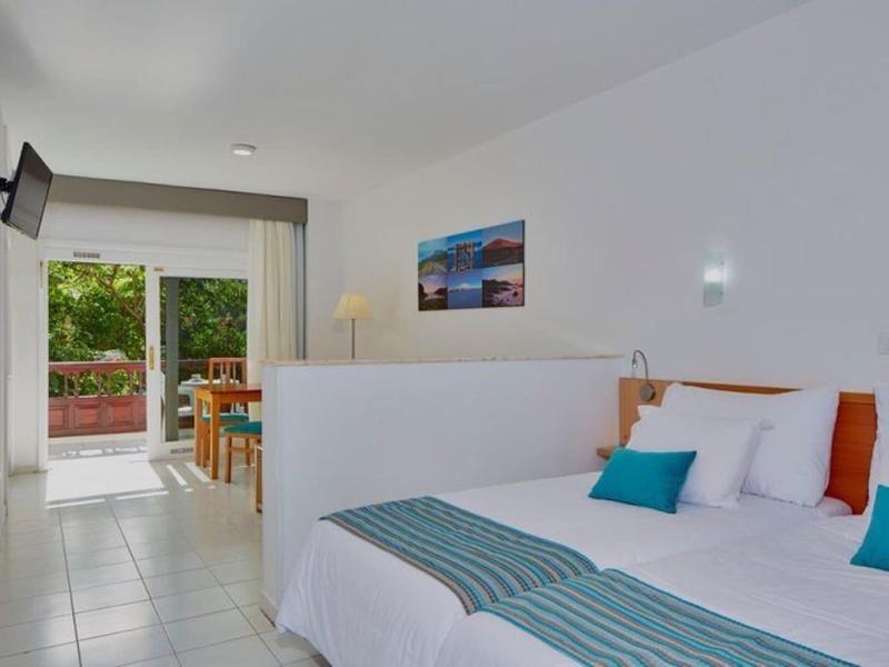 Camera d'hotel luminosa con letto matrimoniale, cuscini blu e zona pranzo vicino a porta di vetro.