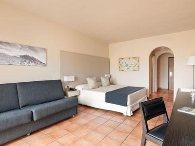 Camera d'hotel luminosa con letto matrimoniale, divano, scrivania e pavimento in terracotta.