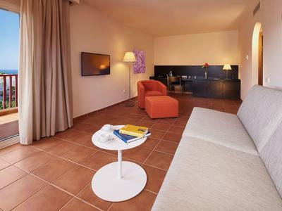 Camera d'hotel accogliente con divano, poltrona, TV, piccolo tavolo bianco e balcone con vista mare.