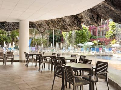 Überdachte Terrasse mit Tischen und Stühlen neben einem Pool und grünen Bäumen.