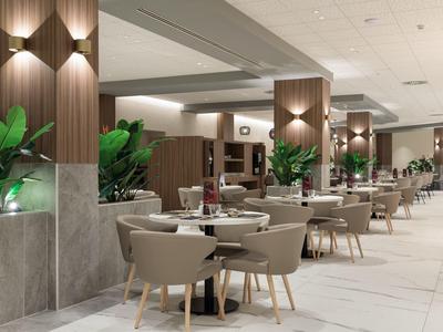Moderner Hotelrestaurantbereich mit runden Tischen, bequemen Stühlen und grünen Pflanzen als Dekoration.