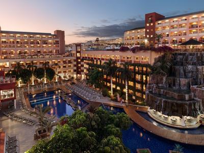 Hotelanlage bei Sonnenuntergang mit beleuchteten Gebäuden, Pool und Wasserfall.