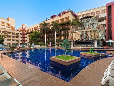 Moderner Hotelpool mit Holzdeck, grünen Pflanzen und Liegestühlen umgeben von mehrstöckigen Gebäuden.