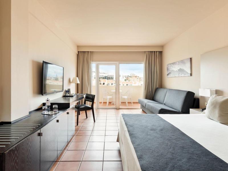 Camera d'albergo luminosa con letto, divano, scrivania e balcone con vista mare.