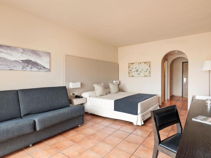 Camera d'hotel luminosa con letto matrimoniale, divano, scrivania e pavimento in terracotta.