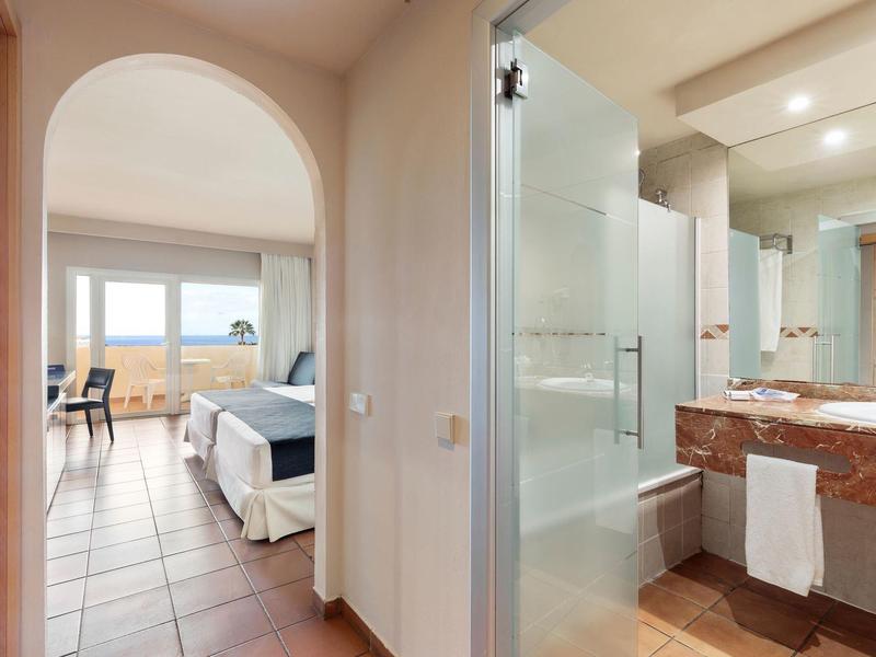 Camera d'hotel luminosa con vista sul mare e bagno adiacente con doccia in vetro.