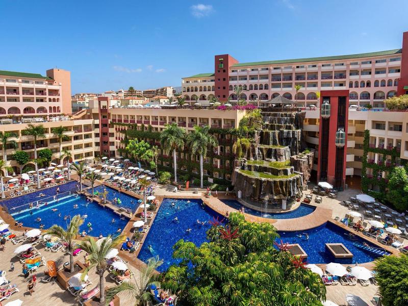 Grande resort con varie piscine, lettini e edifici con vegetazione sotto un cielo sereno.