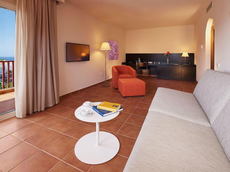 Camera d'hotel accogliente con divano, poltrona, TV, piccolo tavolo bianco e balcone con vista mare.