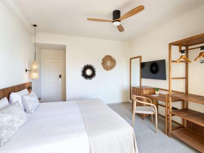 Chambre d'hôtel lumineuse avec lit double, ventilateur, miroir et penderie ouverte en bois.