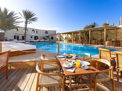 Sonnige Terrasse mit Esstisch, Holzstühlen und klarem Pool vor weißem Gebäude unter blauem Himmel.