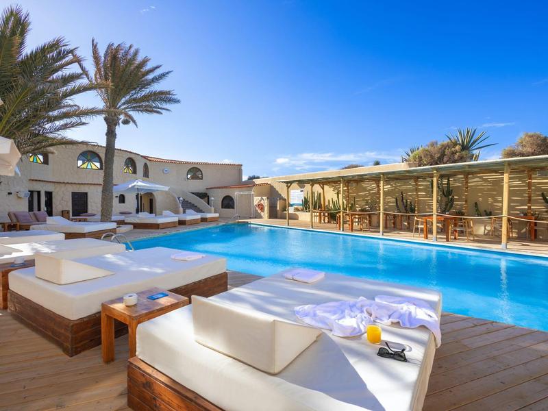 Espace piscine moderne avec transats blancs, palmiers et ciel bleu clair à l'hôtel.