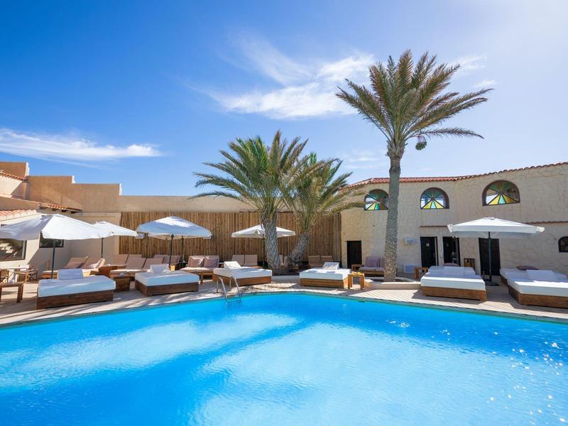 Zone de piscine avec transats, parasols et palmiers sous un ciel bleu.
