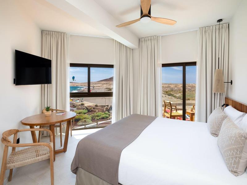 Chambre d'hôtel lumineuse avec lit double, deux chaises, table et vue sur la mer à travers de grandes fenêtres.